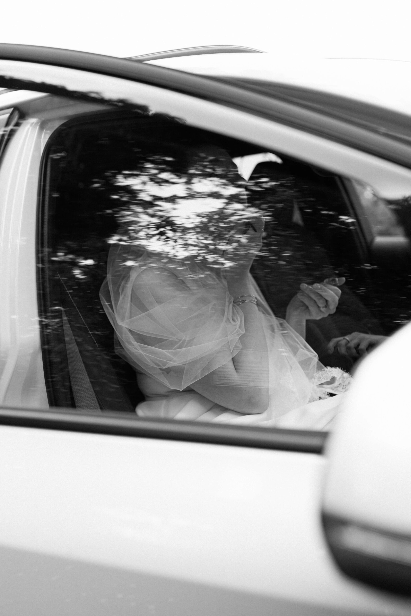 Die Braut sitzt für ein finales Touchup im Auto, in der Scheibe spiegeln sich die Bäume, das Bild ist schwarzweiß und bei der Hochzeitsreportage entstanden